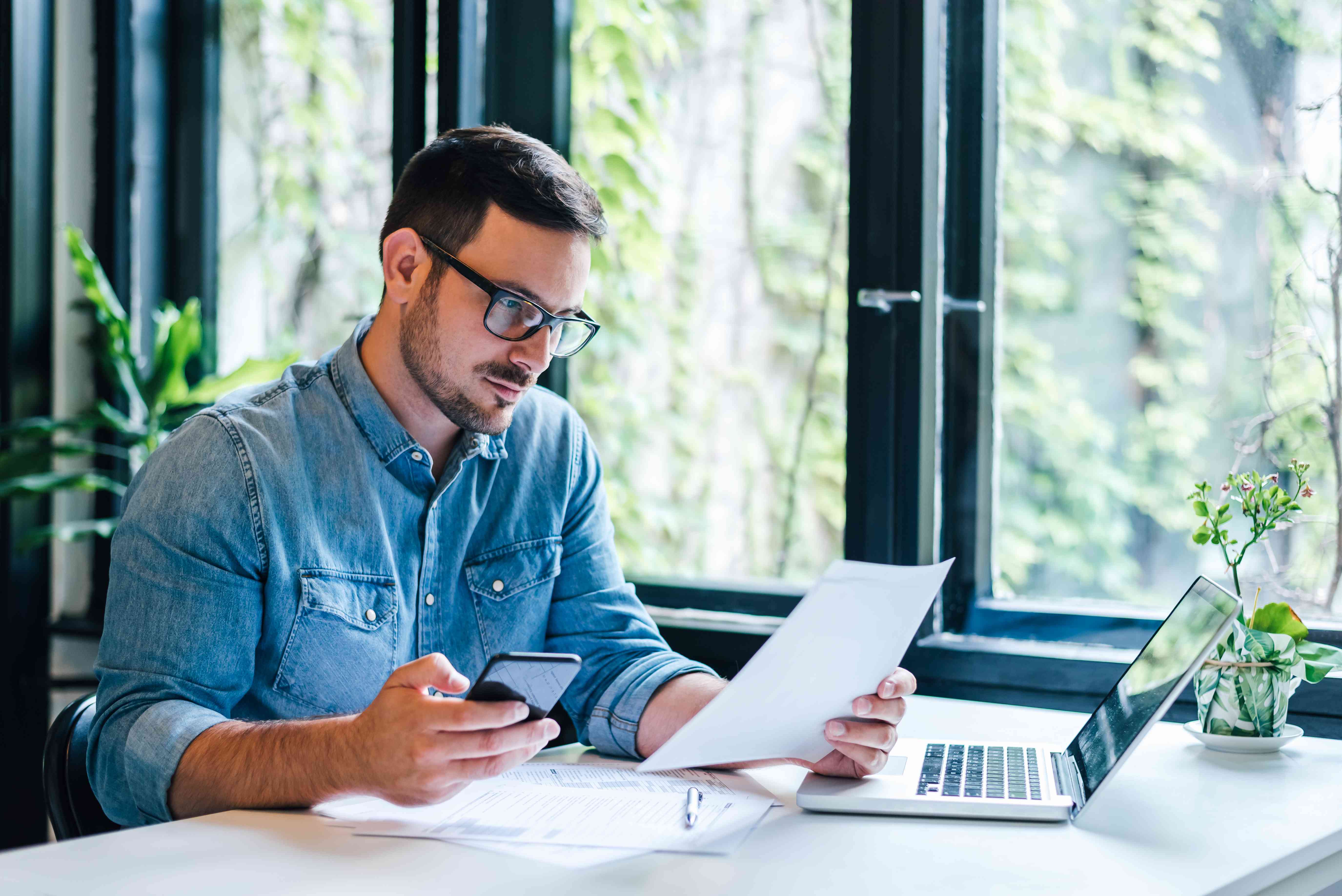 A man sits at a desk with a computer, looking at his phone and some documents.