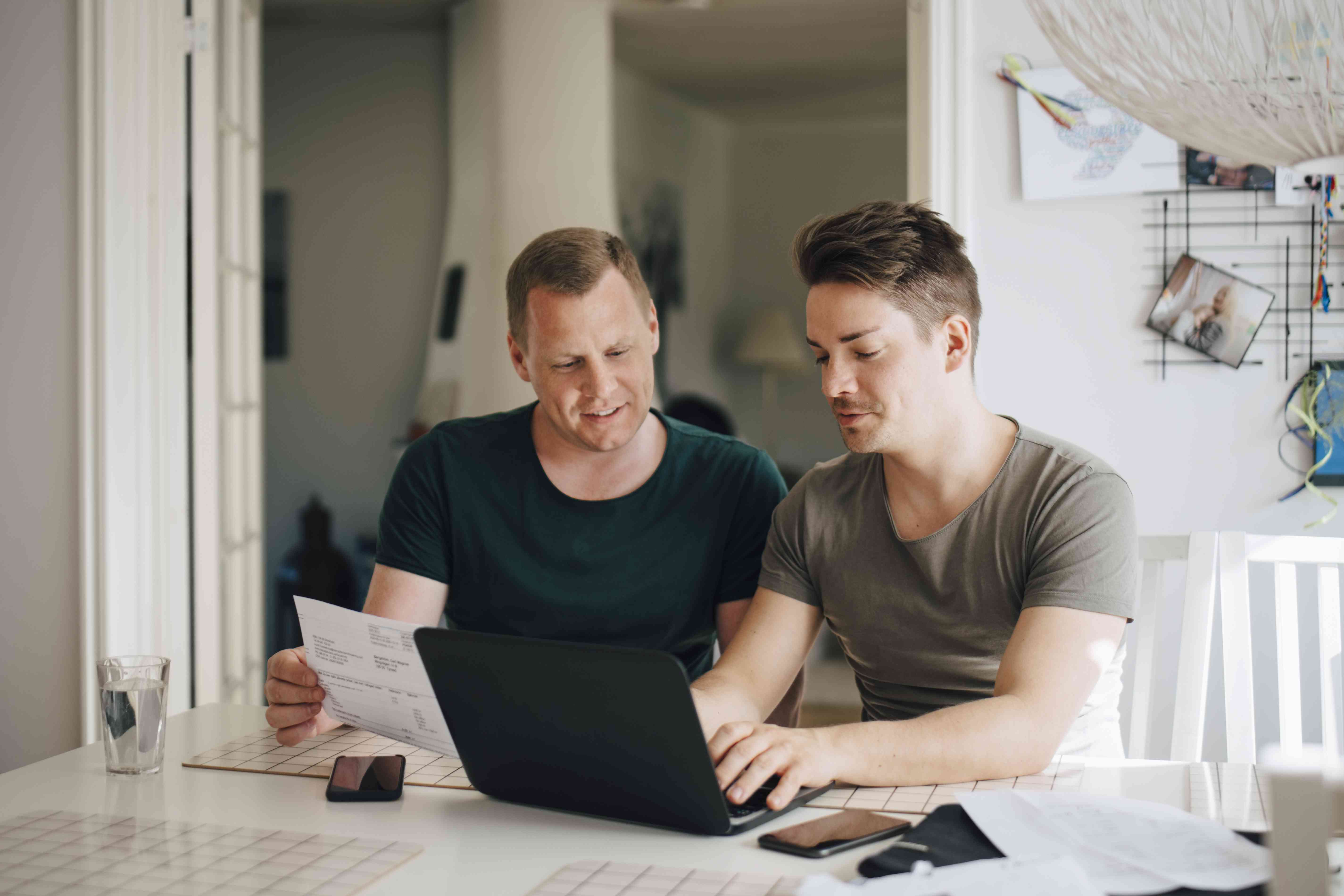 Two men are using a laptop and reviewing documents.