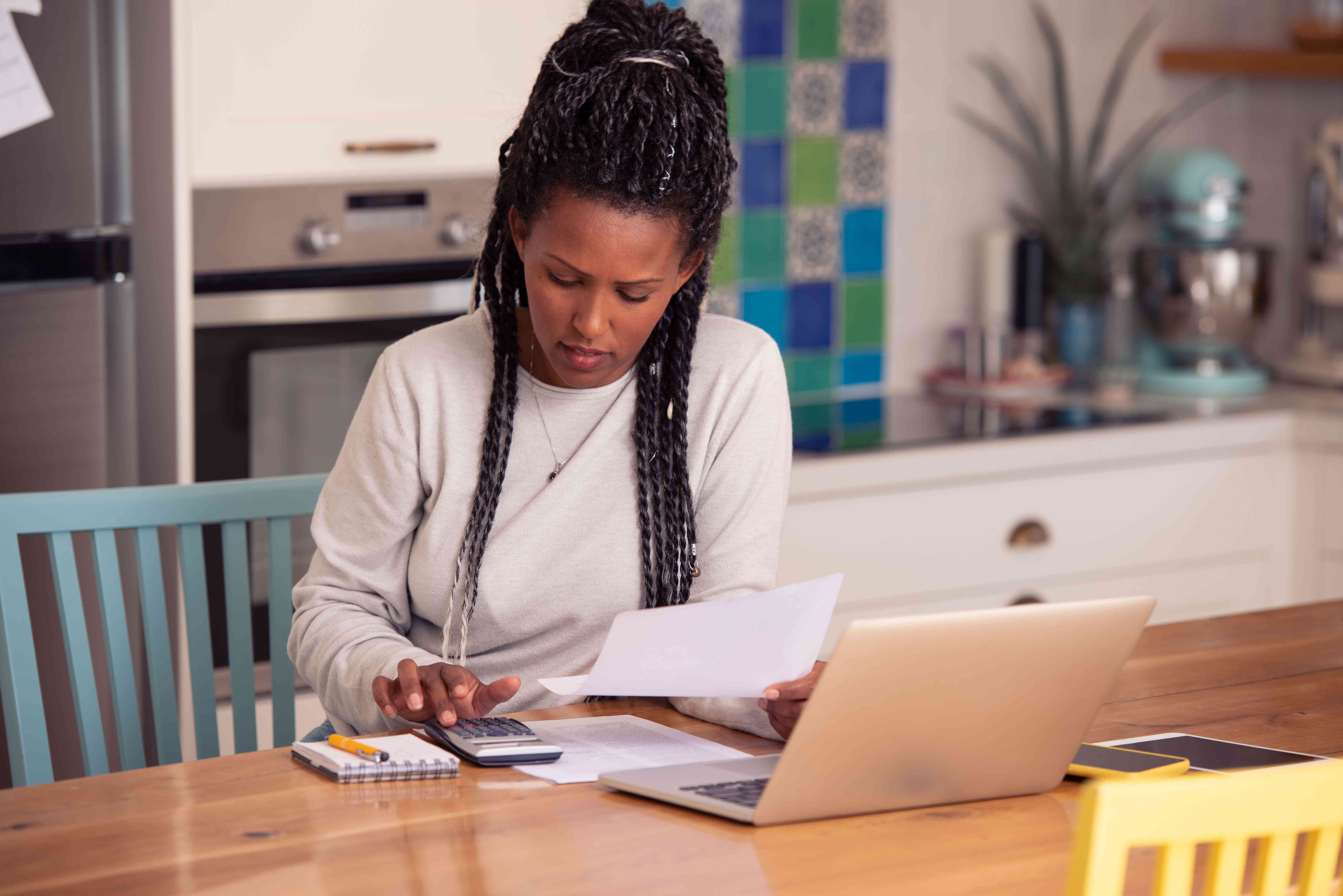 Woman using a calculator with paperwork and a laptop at a kitchen table