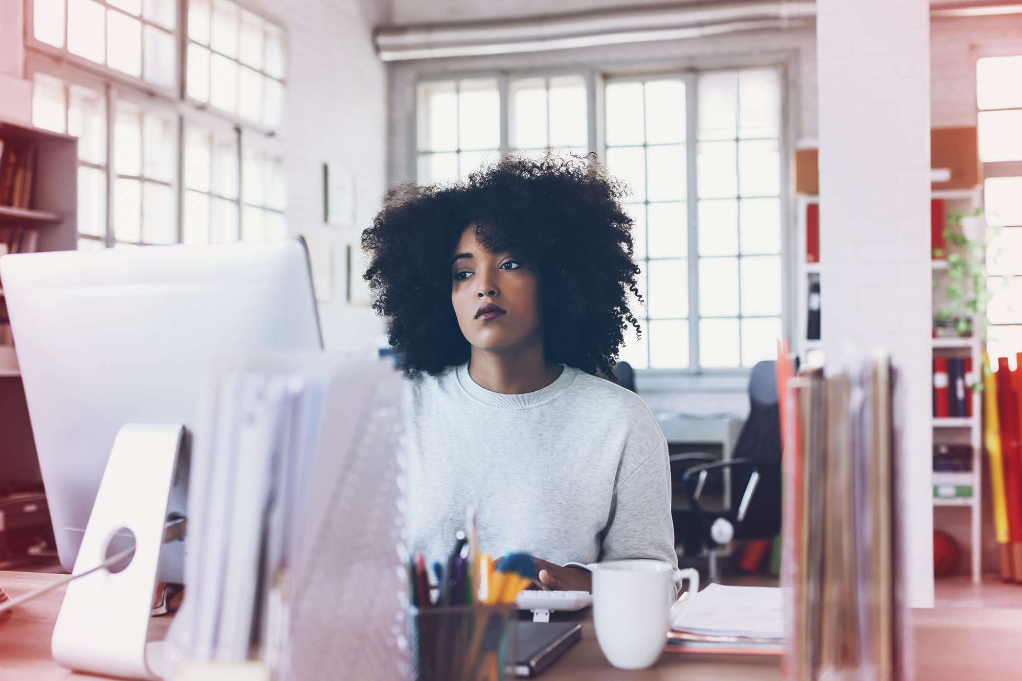 microaggressions - Tired young woman sitting on desk and using computer. Papers and tools, a cup on desk. Tall windows, shelves with folders, office seat on background.