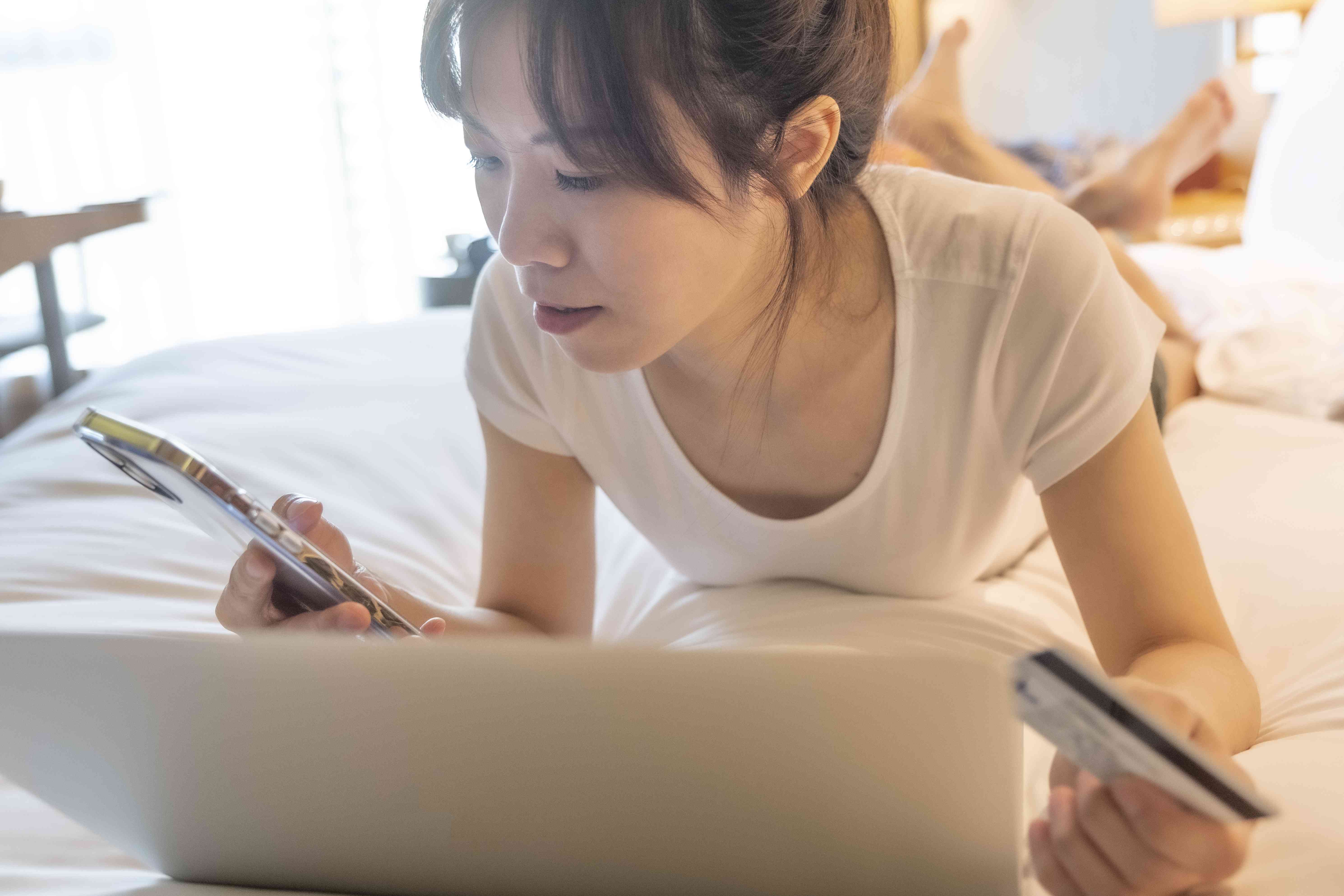 A woman lays on a bed with a laptop and cell phone holding holding a payment card in her hand