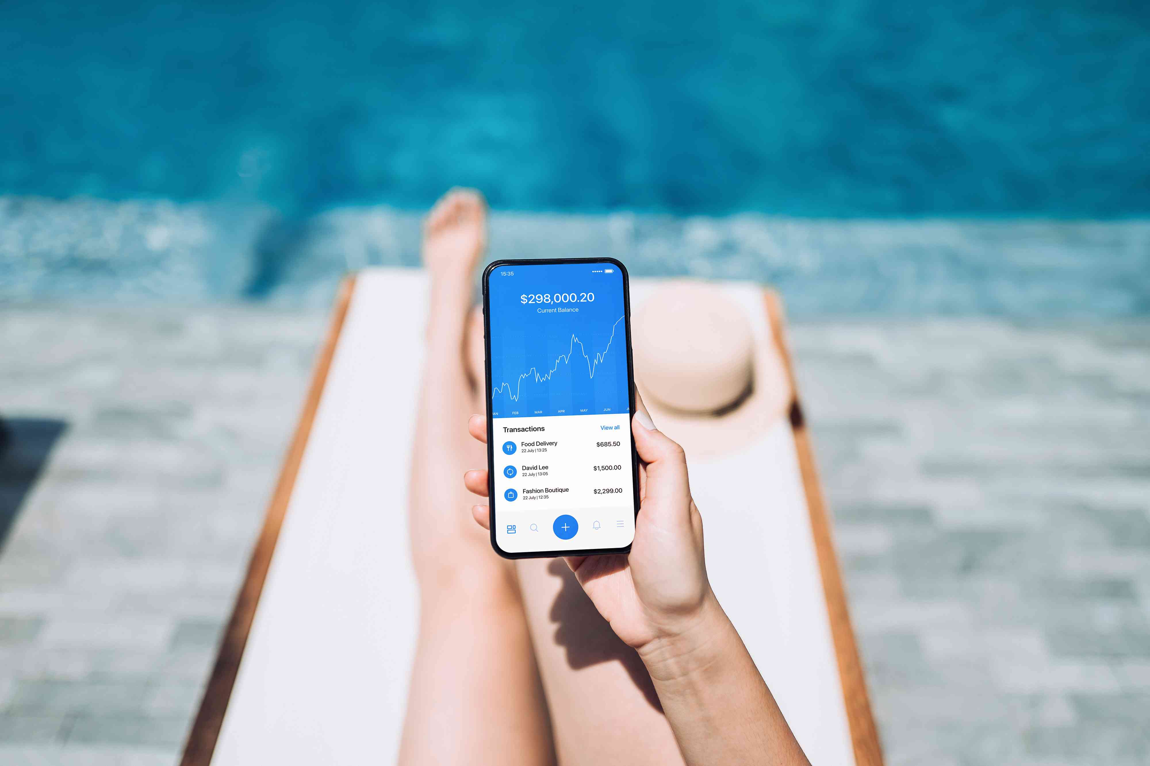 Low section of young woman managing online banking with mobile app on smartphone while enjoying the sun on lounge chair by pool side